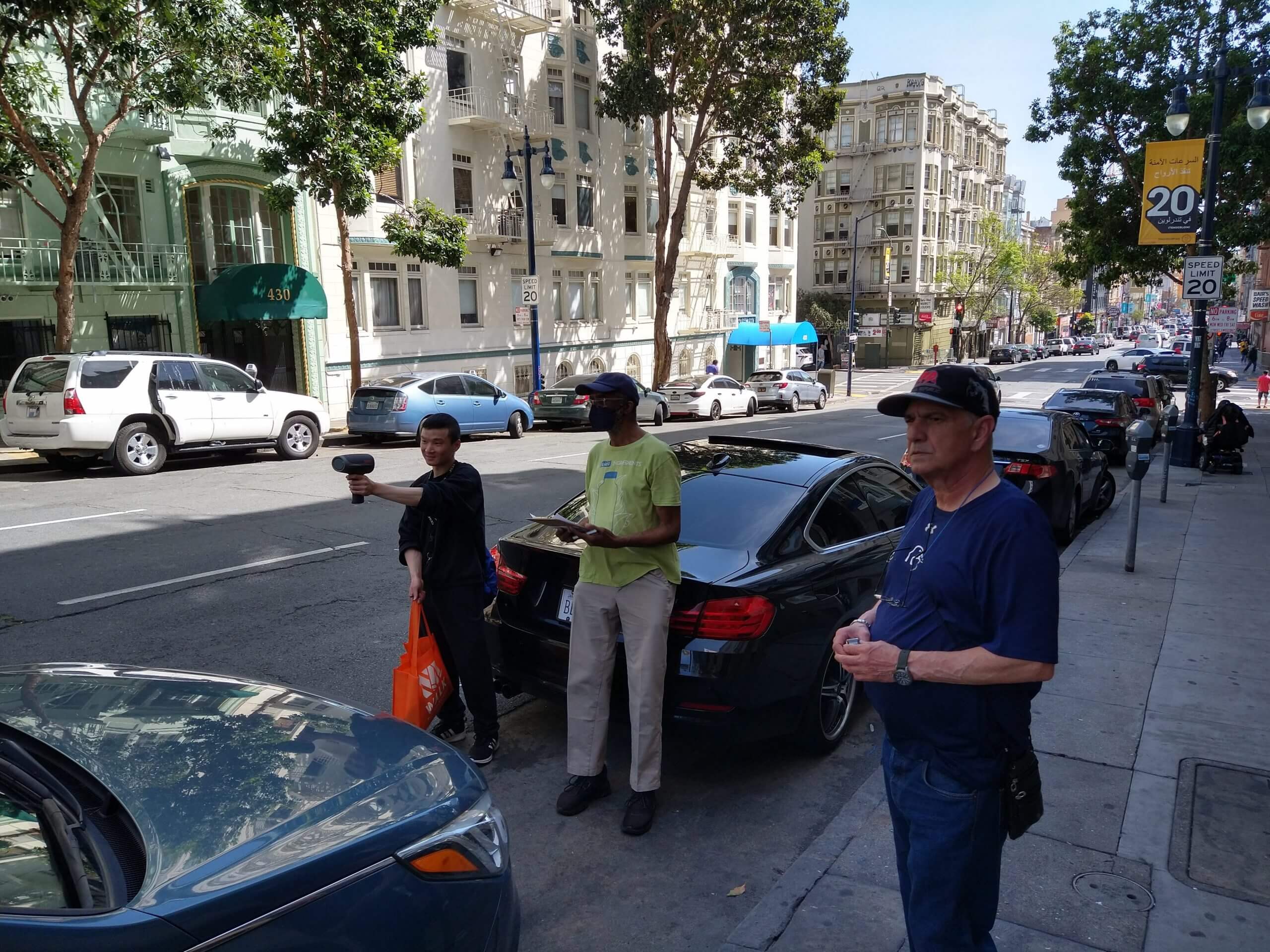 Three volunteers standing on a Tenderloin street. One is holding a speed radar device and another is writing information down in a clipboard.