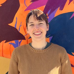 A woman with short hair and a beige sweater smiling in front of a colorful mural
