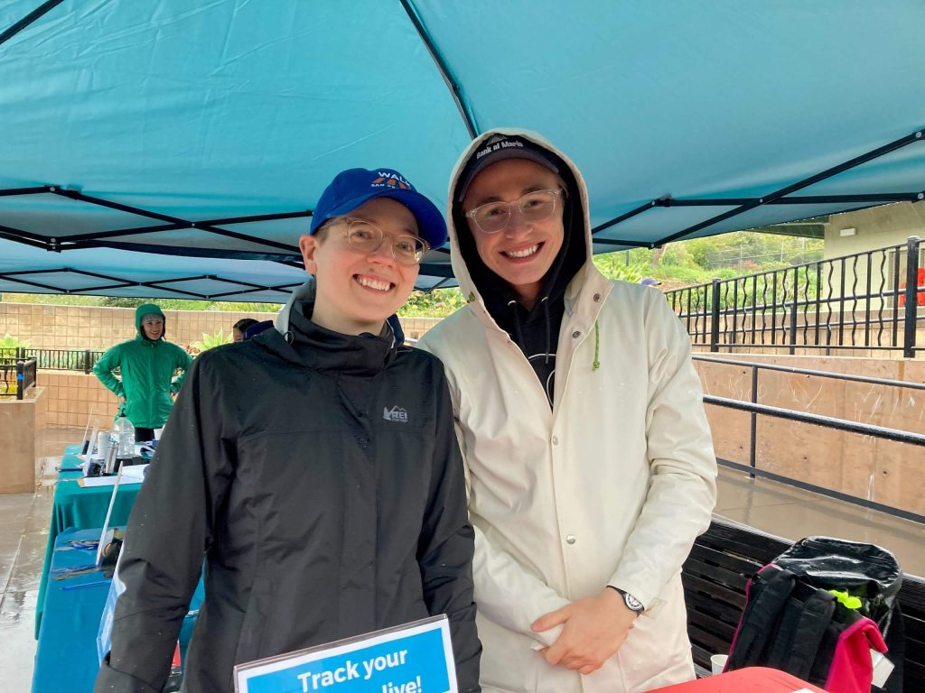 Two individuals pose for a photo under a teal tent.