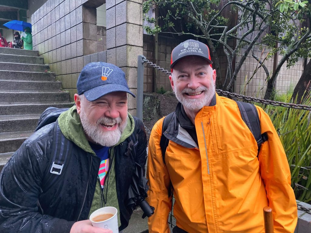 Two bearded individuals wearing gray caps pose for a picture side-by-side.