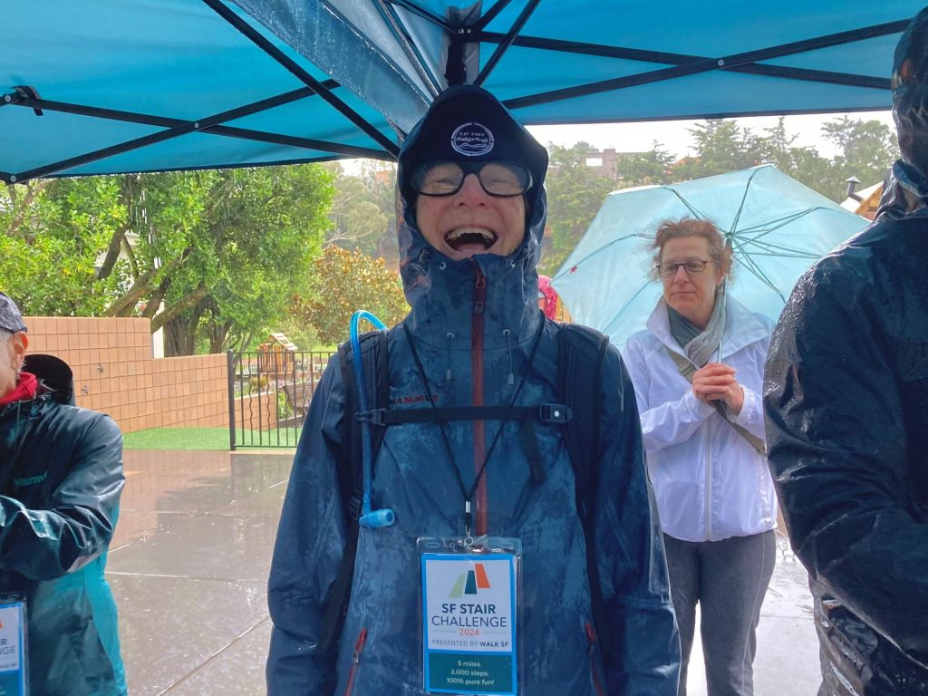 An individual wearing a beanie and rain jacket smiles widely while wearing a Stair Challenge lanyard.